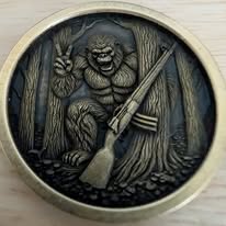 Brass coin engraved with a Bigfoot figure holding a peace sign and a shotgun, standing in front of pine trees